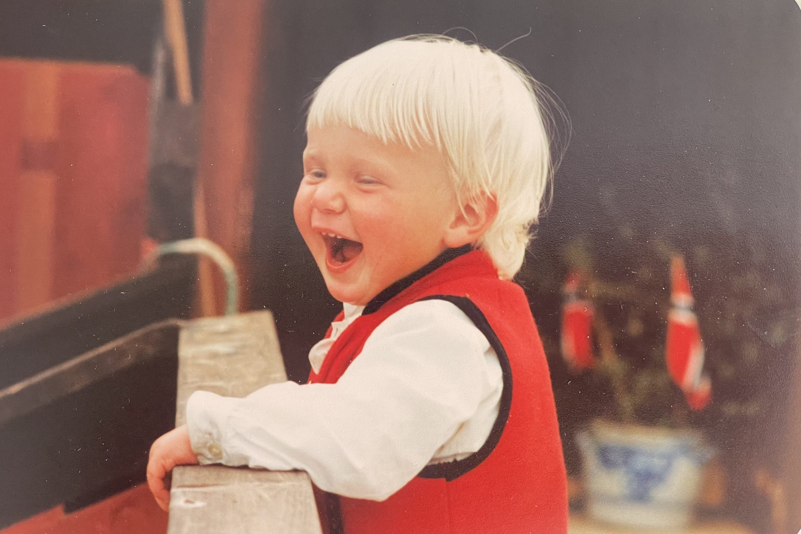 A tiny baby Huld about two years old with bright white hair. She's holding onto a wooden bannister and laughing.