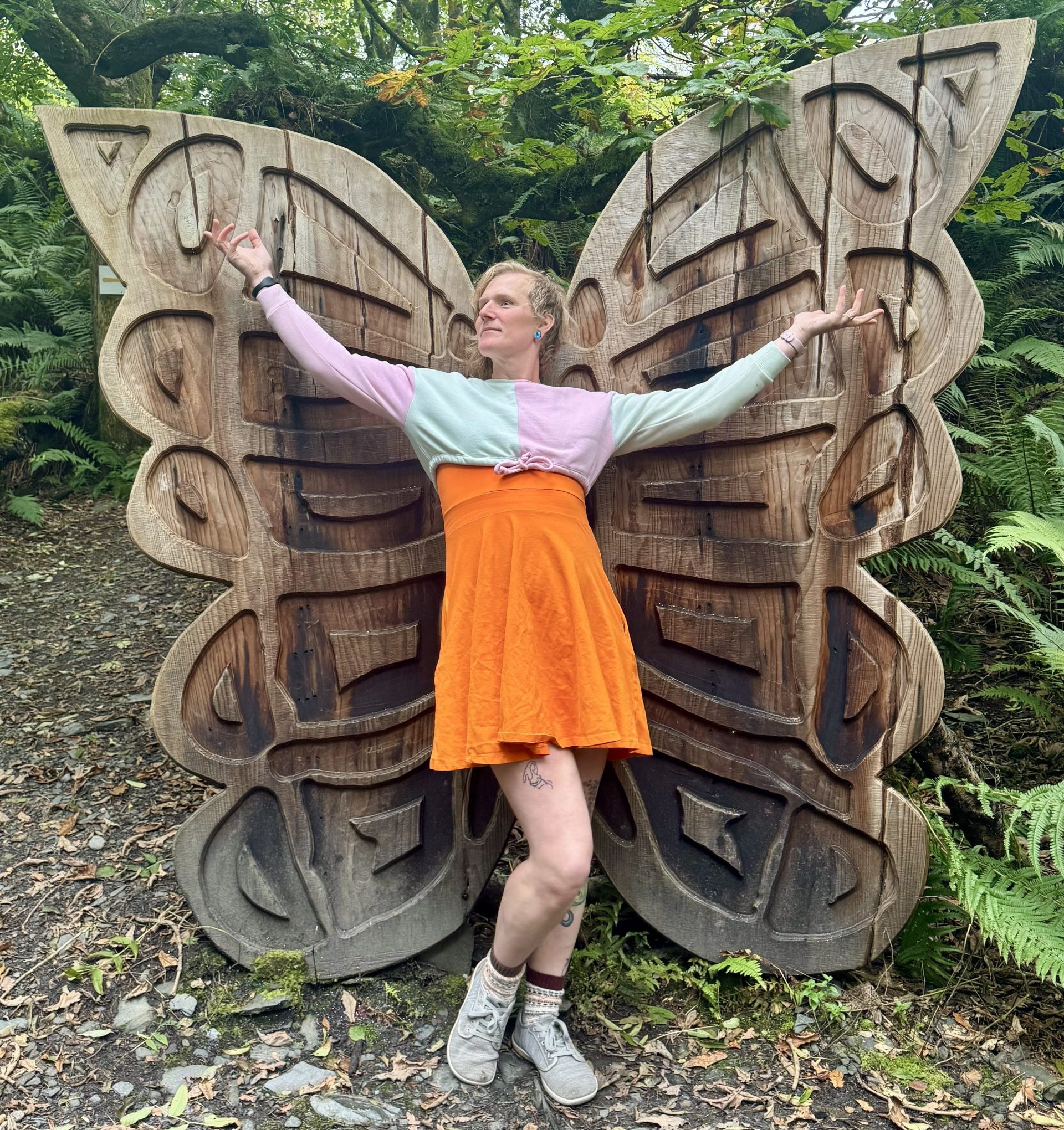 Huld is wearing an orange dress and a pastel crop top. She stands in front of a set of giant wooden butterfly wings with her arms wide